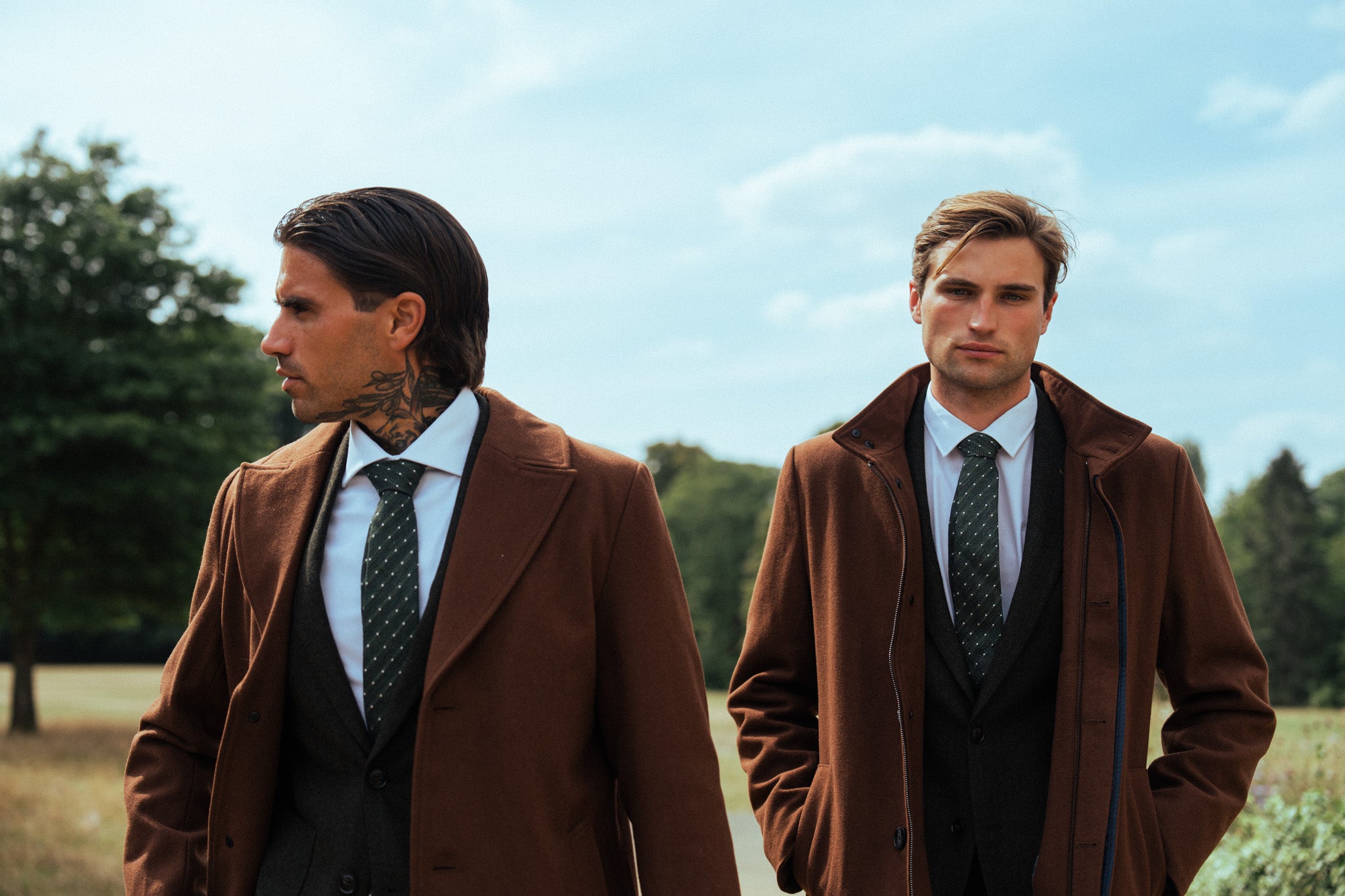Two men in matching Harry Brown London brown coats standing outdoors with trees and blue sky in the background.